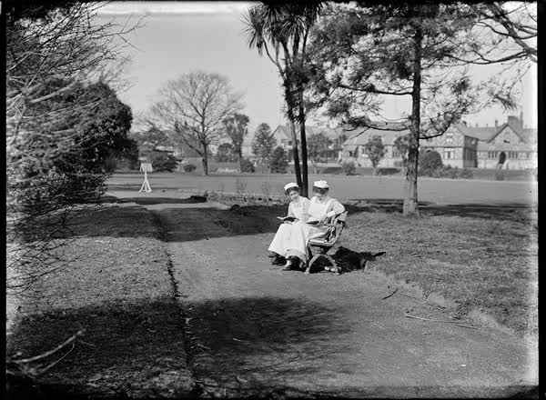 Two women in nurses' uniforms seated on a park bench, 1900