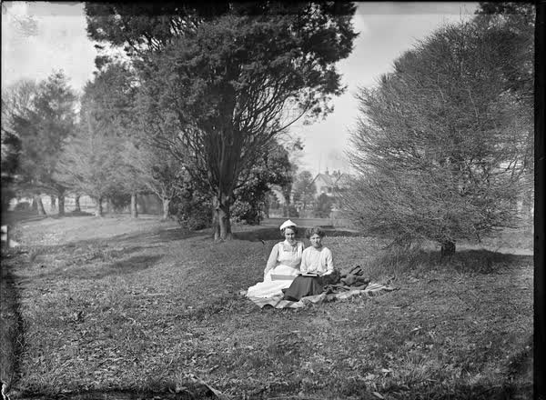 Two women, one in a nurse's uniform, seated on a picnic blanket, 1900