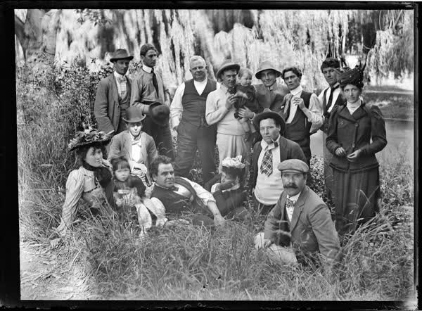 Sixteen men, women and children on the riverbank, 1900